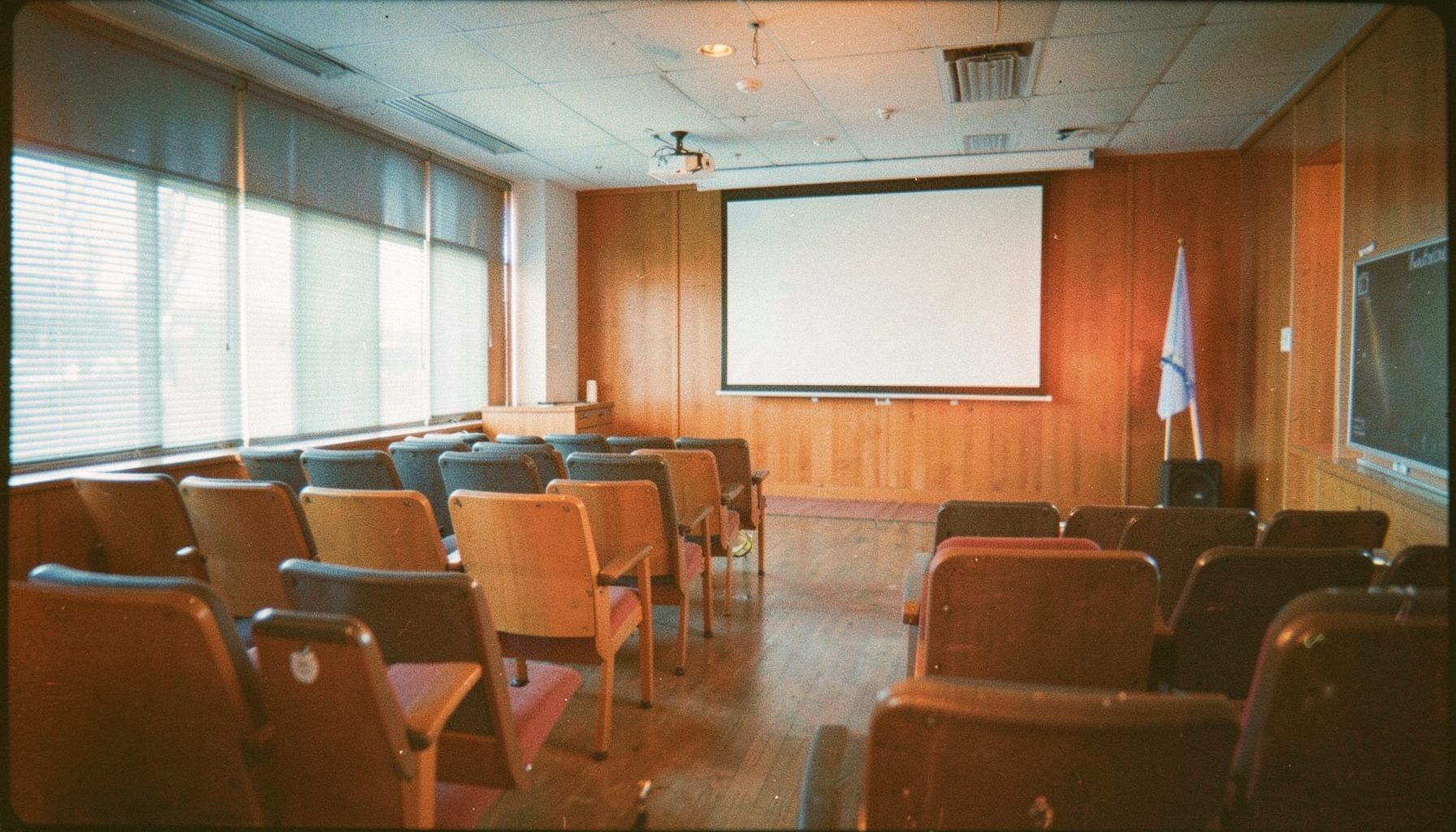 Wide-angle photograph of a conference room with rows of chairs, a projector screen, and soft daylight filtering through blinds, in muted blues and warm wood tones